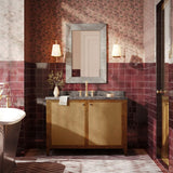 A bathroom vanity setup in a richly decorated room, featuring a wooden cabinet with woven cane doors, a dark stone countertop, a gold faucet, a gray-framed mirror, and deep burgundy tiled walls and floor.